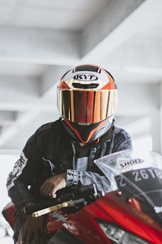 A person wearing a helmet and a denim jacket is seated on a red motorcycle. The helmet has reflective and vibrant colors, including orange and dark tones. The motorcycle has visible brand decals and is parked in an indoor setting with concrete beams visible in the background.