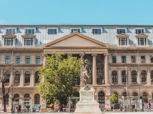 A large, historic building with an inscription reading 'UNIVERSITATEA' on its pediment. The façade is adorned with arched windows and decorative columns. There is a prominent statue in the foreground on a plinth, surrounded by greenery and some people walking nearby. The walls of the building feature graffiti at the bottom.