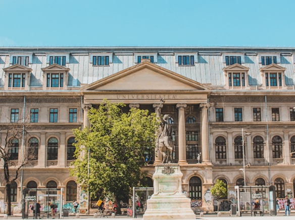 A large, historic building with an inscription reading 'UNIVERSITATEA' on its pediment. The façade is adorned with arched windows and decorative columns. There is a prominent statue in the foreground on a plinth, surrounded by greenery and some people walking nearby. The walls of the building feature graffiti at the bottom.