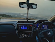The interior of a car is visible with the dashboard prominently featuring a digital display and controls. The steering wheel with a recognizable logo is on the right side. Through the windshield, an open landscape can be seen, including a river and distant hills under a cloudy sky.