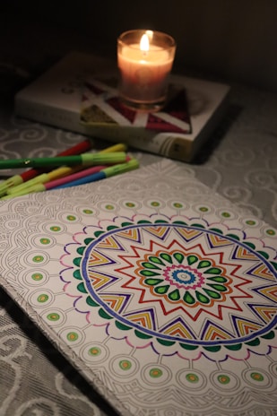 A candle lit on top of a book casts a warm glow over colored markers and a partially colored mandala design on a table with a textured tablecloth.