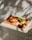 A slice of pizza with a golden-brown crust, topped with melted cheese, tomato sauce, cherry tomatoes, and a fresh basil leaf, sits on a wooden board. The background features a tiled wall with dappled sunlight and shadows creating an artistic effect.