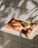A slice of pizza with a golden-brown crust, topped with melted cheese, tomato sauce, cherry tomatoes, and a fresh basil leaf, sits on a wooden board. The background features a tiled wall with dappled sunlight and shadows creating an artistic effect.