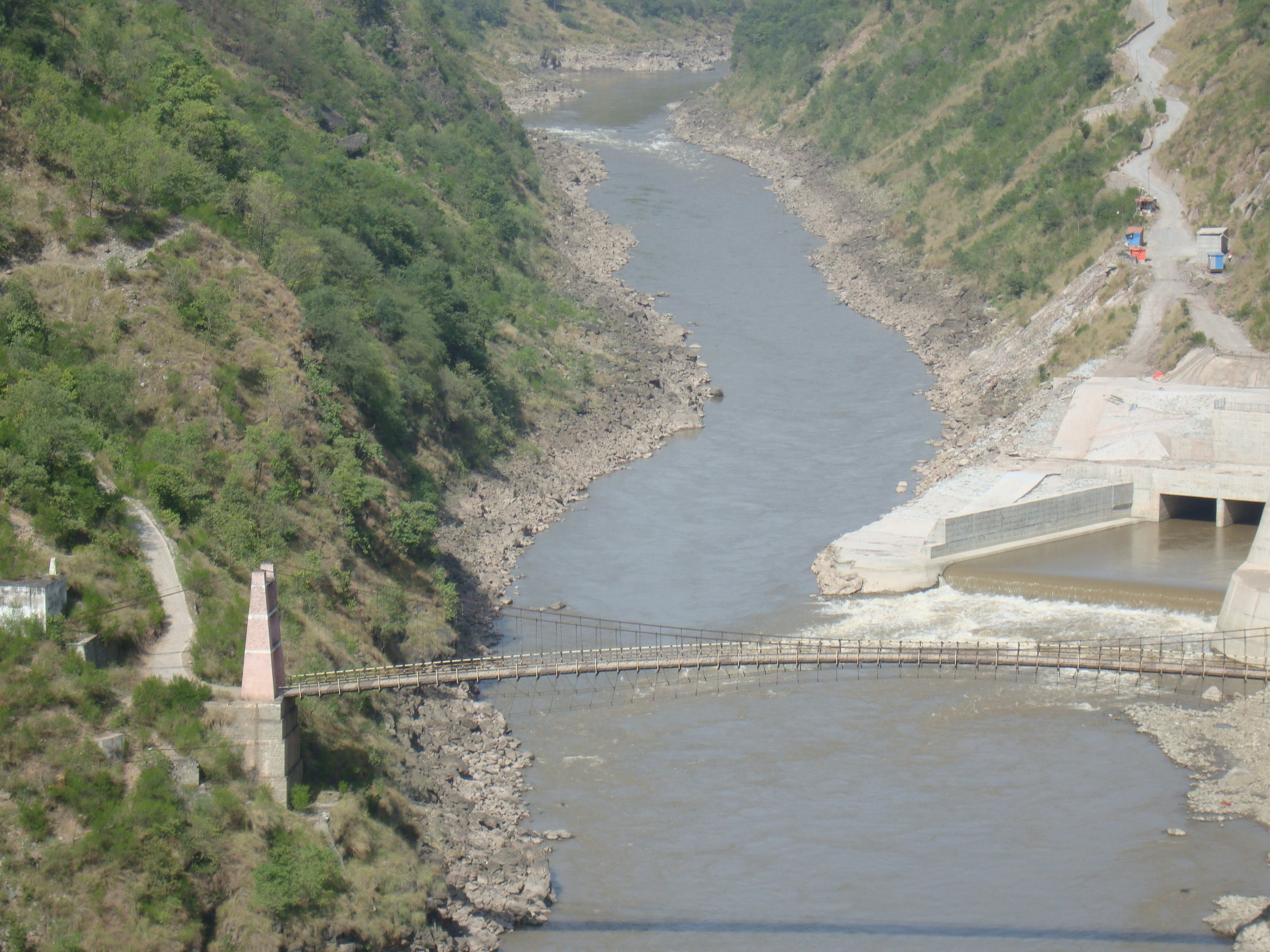 A narrow suspension bridge spans a winding river through a rocky canyon, with a dam and a distant road along the riverbank.