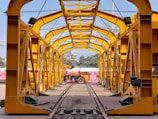 A yellow metal structure resembling a tunnel or framework, with a clear view towards an outdoor area including trees and a partially visible building. A vehicle, possibly a construction machine, is positioned at the end of the framework.