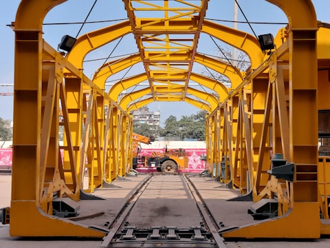 A yellow metal structure resembling a tunnel or framework, with a clear view towards an outdoor area including trees and a partially visible building. A vehicle, possibly a construction machine, is positioned at the end of the framework.