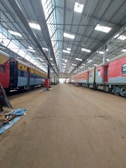 Inside a spacious train maintenance workshop, two trains are parked parallel to each other under a high, metal-framed ceiling with skylights. The floor is dusty and cluttered with debris in some areas.