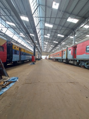 Inside a spacious train maintenance workshop, two trains are parked parallel to each other under a high, metal-framed ceiling with skylights. The floor is dusty and cluttered with debris in some areas.