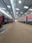 Inside a spacious train maintenance workshop, two trains are parked parallel to each other under a high, metal-framed ceiling with skylights. The floor is dusty and cluttered with debris in some areas.