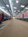 Inside a spacious train maintenance workshop, two trains are parked parallel to each other under a high, metal-framed ceiling with skylights. The floor is dusty and cluttered with debris in some areas.