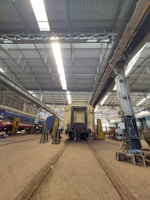 A large industrial train workshop with high ceilings, showcasing a train car undergoing maintenance. The space is organized with various tools and equipment scattered around, along with workers in the background attending to their tasks. The lighting is bright, with overhead lights reflecting off the metallic surfaces.