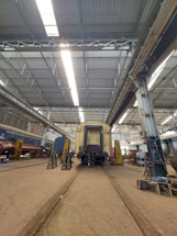 A large industrial train workshop with high ceilings, showcasing a train car undergoing maintenance. The space is organized with various tools and equipment scattered around, along with workers in the background attending to their tasks. The lighting is bright, with overhead lights reflecting off the metallic surfaces.