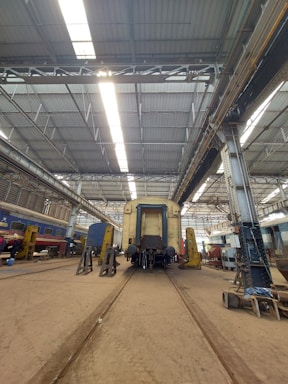 A large industrial train workshop with high ceilings, showcasing a train car undergoing maintenance. The space is organized with various tools and equipment scattered around, along with workers in the background attending to their tasks. The lighting is bright, with overhead lights reflecting off the metallic surfaces.