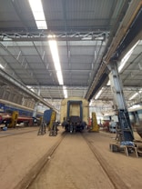 A large industrial train workshop with high ceilings, showcasing a train car undergoing maintenance. The space is organized with various tools and equipment scattered around, along with workers in the background attending to their tasks. The lighting is bright, with overhead lights reflecting off the metallic surfaces.