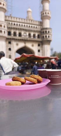 A pink plate with biscuits is placed on a metallic surface, next to a brown cup printed with text. In the background, a historic monument with arches and minarets looms atop a busy outdoor market scene. People can be seen moving about, and a red canopy covers the stall.