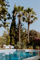 A tranquil outdoor scene featuring a swimming pool surrounded by lush greenery and tall palm trees. White lounge chairs are placed by the poolside, offering a relaxing spot under the clear blue sky. The water in the pool reflects the surrounding foliage.