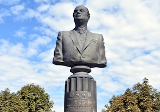 A large bronze bust of a man dressed in a formal suit stands prominently against a backdrop of a partly cloudy blue sky. The bust is mounted on a pedestal with inscriptions and various medals. Surrounding the base of the pedestal are green trees, adding to the natural setting.
