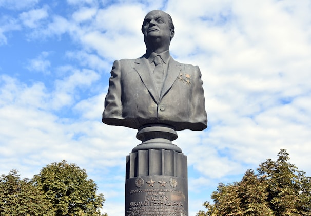 A large bronze bust of a man dressed in a formal suit stands prominently against a backdrop of a partly cloudy blue sky. The bust is mounted on a pedestal with inscriptions and various medals. Surrounding the base of the pedestal are green trees, adding to the natural setting.
