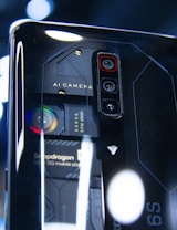 A close-up view of a smartphone's back panel showcasing its triple camera setup and other technical components. The text 'AI Camera' and 'Snapdragon' can be seen on the phone, along with visible lenses and intricate design patterns in a futuristic style.