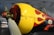 A close-up of a vintage aircraft's propeller area, featuring a yellow and black paint scheme with red flames. The aircraft is inside a hangar with industrial lighting. The propeller is metallic, and rivets are visible on the fuselage.