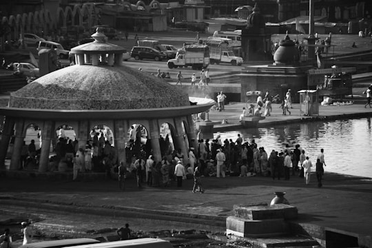 A large dome-shaped pavilion by a waterfront with a crowd of people gathered underneath and around it. The area is bustling, with people walking and engaging in various activities. Several vehicles, including buses and cars, are parked in the background, and there are architectural structures lining the scene.