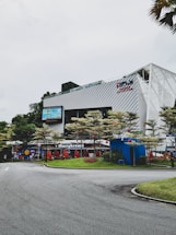 A commercial building with modern architecture featuring metal paneling and large glass windows. There is a restaurant named 'Marrybrown' on the ground floor, surrounded by well-maintained landscaping and trees adorned with red lanterns. The sky is overcast, and a curved road leads into the area.