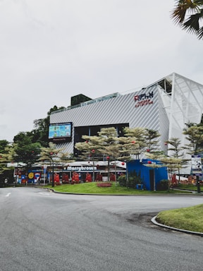 A commercial building with modern architecture featuring metal paneling and large glass windows. There is a restaurant named 'Marrybrown' on the ground floor, surrounded by well-maintained landscaping and trees adorned with red lanterns. The sky is overcast, and a curved road leads into the area.