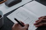 A person writes in a journal on a gray felt surface. The journal page includes sections for checking mood options like 'happy' or 'worried', and areas for writing down intentions and things to be grateful for. A cup of coffee sits in the corner, and a pen case is partially visible.