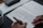 A person writes in a journal on a gray felt surface. The journal page includes sections for checking mood options like 'happy' or 'worried', and areas for writing down intentions and things to be grateful for. A cup of coffee sits in the corner, and a pen case is partially visible.