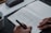 A person writes in a journal on a gray felt surface. The journal page includes sections for checking mood options like 'happy' or 'worried', and areas for writing down intentions and things to be grateful for. A cup of coffee sits in the corner, and a pen case is partially visible.