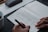 A person writes in a journal on a gray felt surface. The journal page includes sections for checking mood options like 'happy' or 'worried', and areas for writing down intentions and things to be grateful for. A cup of coffee sits in the corner, and a pen case is partially visible.