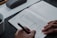 A person writes in a journal on a gray felt surface. The journal page includes sections for checking mood options like 'happy' or 'worried', and areas for writing down intentions and things to be grateful for. A cup of coffee sits in the corner, and a pen case is partially visible.