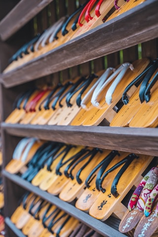 Rows of traditional wooden sandals with straps are neatly arranged on shelves. The sandals feature various colors of straps, including black, red, and white.