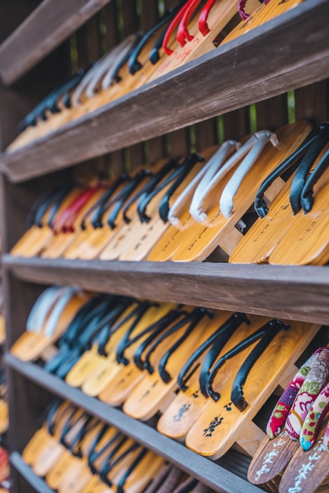 Rows of traditional wooden sandals with straps are neatly arranged on shelves. The sandals feature various colors of straps, including black, red, and white.