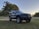 A dark gray SUV is parked on a grassy area with a tree-lined background and a clear sky. The vehicle is equipped with off-road tires, chrome rims, and a roof rack with additional lighting.