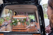 A wicker casket adorned with assorted flowers, placed inside a hearse. The flowers include purple, pink, and white blooms surrounded by green foliage. A person is partially visible on the right side.