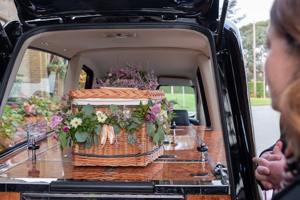 A wicker casket adorned with assorted flowers, placed inside a hearse. The flowers include purple, pink, and white blooms surrounded by green foliage. A person is partially visible on the right side.
