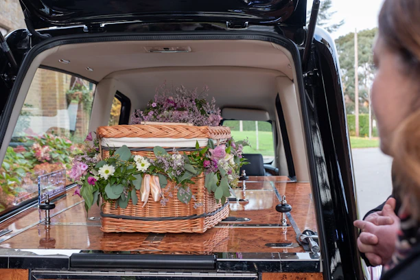 A wicker casket adorned with assorted flowers, placed inside a hearse. The flowers include purple, pink, and white blooms surrounded by green foliage. A person is partially visible on the right side.