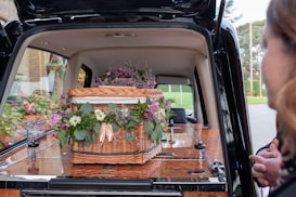 A wicker casket adorned with assorted flowers, placed inside a hearse. The flowers include purple, pink, and white blooms surrounded by green foliage. A person is partially visible on the right side.