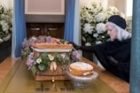 A woman dressed in black with gray hair and gloves is touching a wicker coffin adorned with purple and white flowers. A cake is placed on a glass stand beside it. The setting appears to be a somber indoor space with large bouquets of white flowers in the background.