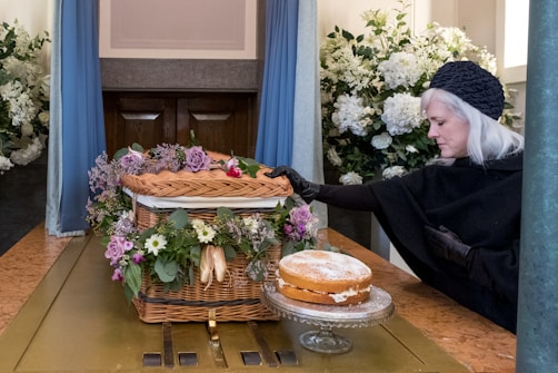 A woman dressed in black with gray hair and gloves is touching a wicker coffin adorned with purple and white flowers. A cake is placed on a glass stand beside it. The setting appears to be a somber indoor space with large bouquets of white flowers in the background.