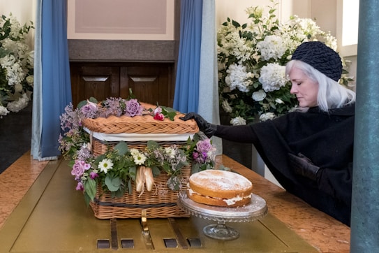 A woman dressed in black with gray hair and gloves is touching a wicker coffin adorned with purple and white flowers. A cake is placed on a glass stand beside it. The setting appears to be a somber indoor space with large bouquets of white flowers in the background.