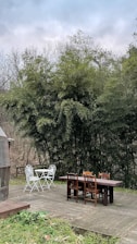A wooden deck set in a natural environment with two sets of outdoor furniture: a white metal table with two chairs and a larger wooden table with four chairs. Surrounding the deck are dense bamboo plants, and there's a clear, octagonal greenhouse structure on the left.