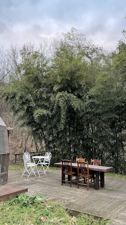 A wooden deck set in a natural environment with two sets of outdoor furniture: a white metal table with two chairs and a larger wooden table with four chairs. Surrounding the deck are dense bamboo plants, and there's a clear, octagonal greenhouse structure on the left.