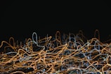 A chaotic array of swirling and looping lines in various colors, mainly orange and blue, set against a dark background. The patterns resemble light trails captured with long exposure.
