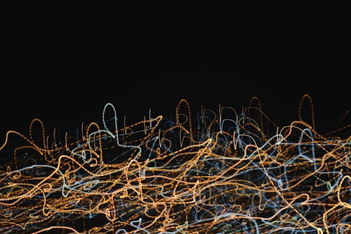 A chaotic array of swirling and looping lines in various colors, mainly orange and blue, set against a dark background. The patterns resemble light trails captured with long exposure.