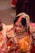 A couple dressed in traditional Indian attire is engaged in a marriage ceremony. The woman, adorned with intricate jewelry and a beautifully embroidered red and orange sari, is being held by the man. The man is wearing a yellow traditional outfit with pink beads around his neck. They are surrounded by celebratory elements, including a pot with flowers.