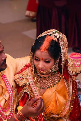A couple dressed in traditional Indian attire is engaged in a marriage ceremony. The woman, adorned with intricate jewelry and a beautifully embroidered red and orange sari, is being held by the man. The man is wearing a yellow traditional outfit with pink beads around his neck. They are surrounded by celebratory elements, including a pot with flowers.