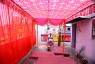 A brightly decorated canopy area with pink and red fabric draped from above. There are flowers and garlands hanging from the canopy. Two individuals, one sitting and one standing, are present in the space. The setting appears to be part of a celebratory or ceremonial event. The ground is covered with striped mats and there are various objects including a red pot and plants.