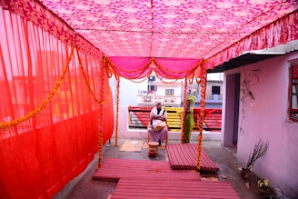 A brightly decorated canopy area with pink and red fabric draped from above. There are flowers and garlands hanging from the canopy. Two individuals, one sitting and one standing, are present in the space. The setting appears to be part of a celebratory or ceremonial event. The ground is covered with striped mats and there are various objects including a red pot and plants.